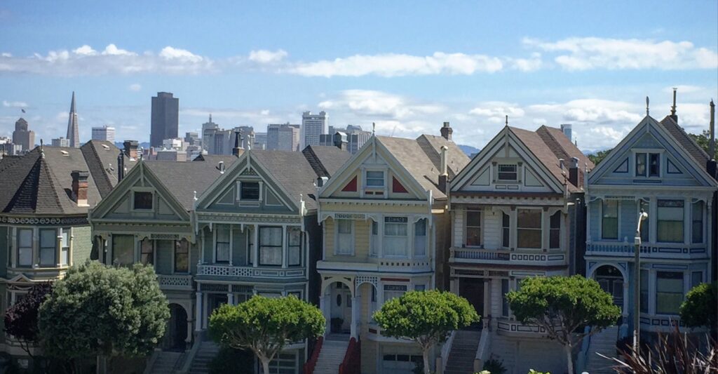 A row of colorful Victorian homes known as the Painted Ladies stands in the foreground, framed by green trees in Alamo Square Park. Behind them, the San Francisco skyline rises under a bright blue sky dotted with white clouds, with the Transamerica Pyramid clearly visible.