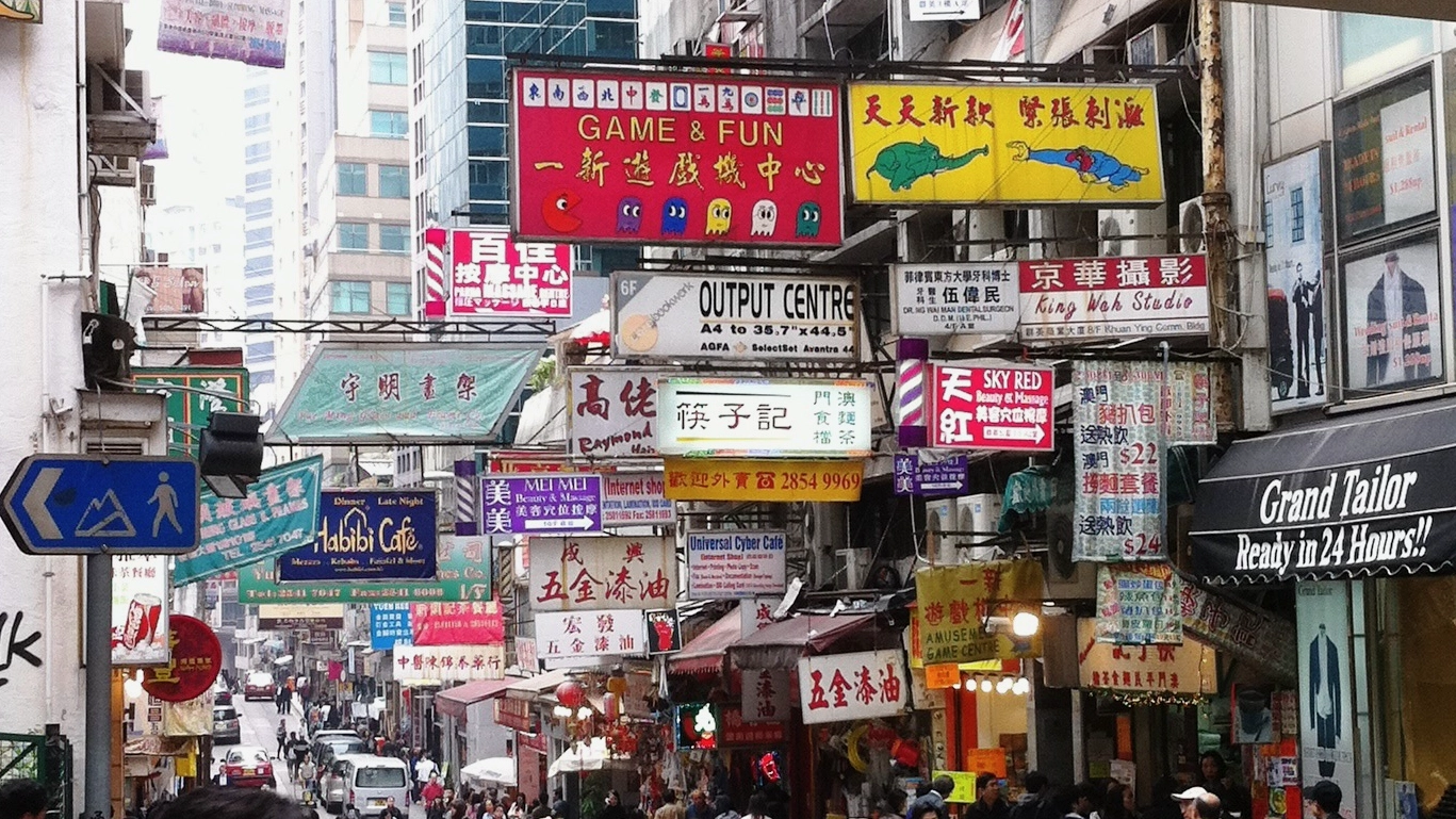 Busy Hong Kong street lined with colorful Chinese and English business signs, shops, pedestrians, and high-rise buildings in a dense urban district.