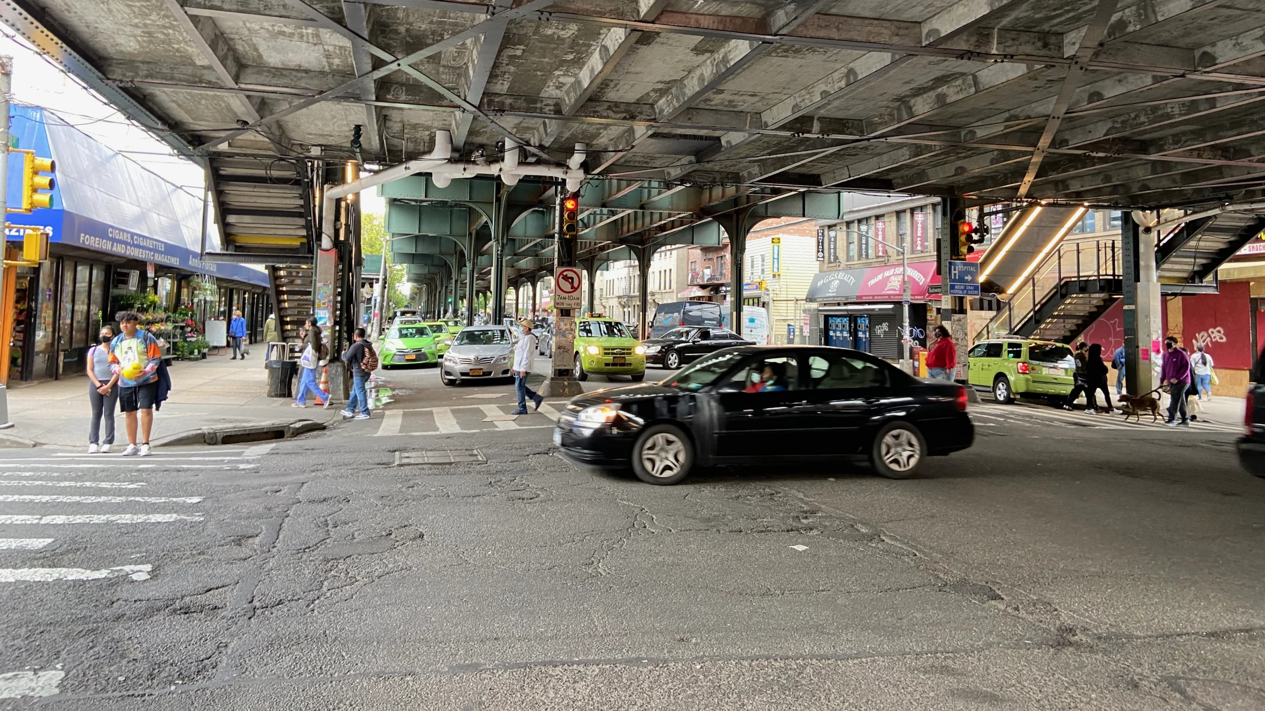 Street-level view beneath elevated subway tracks in Queens, New York, with pedestrians crossing an intersection, cars passing through traffic, and neighborhood storefronts lining the block.