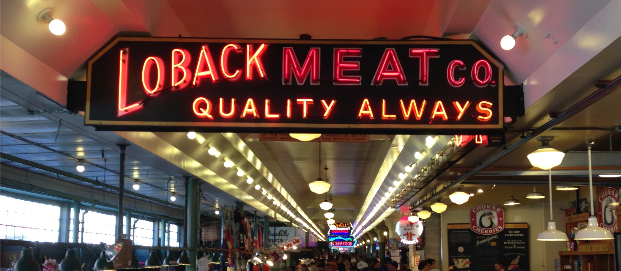 A neon Loback Meat Co. sign hangs above a busy indoor public market corridor filled with shoppers, vendor stalls, and hanging lights. The market features food vendors, flowers, specialty products, and a bustling atmosphere.