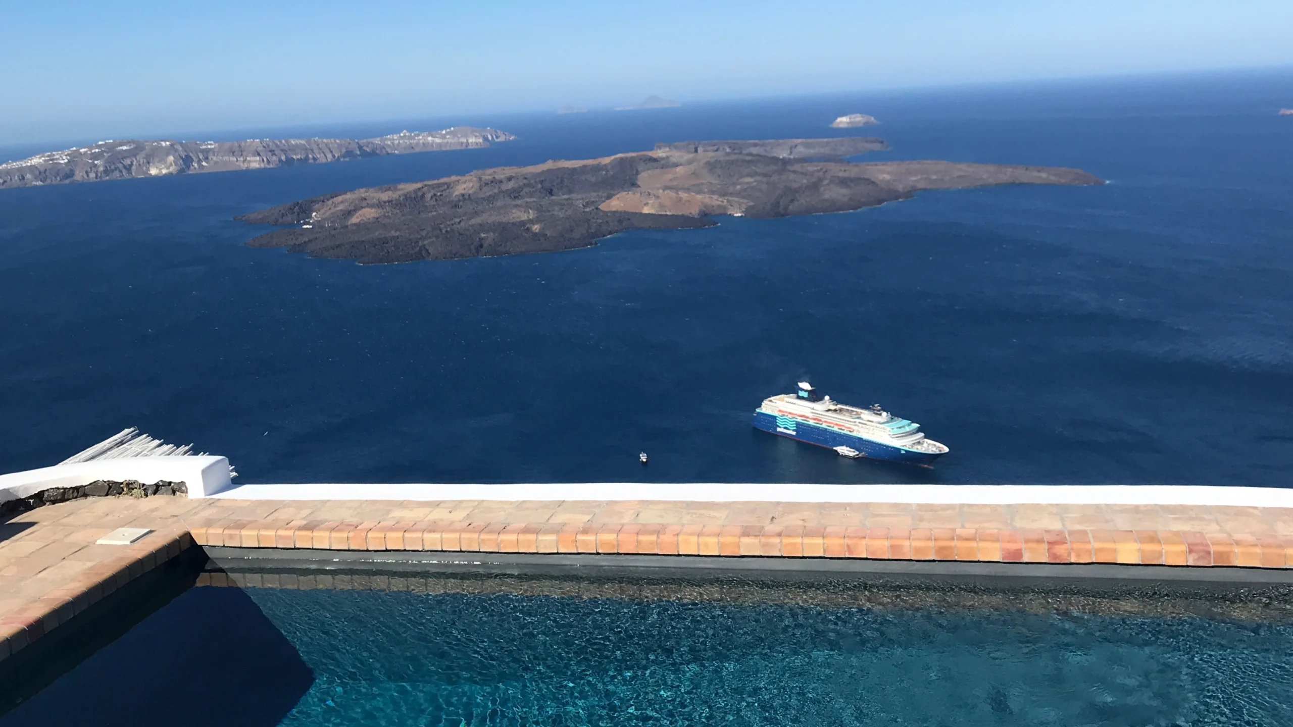 Infinity pool overlooking the Santorini caldera with a cruise ship anchored in the Aegean Sea and volcanic islands in the background.