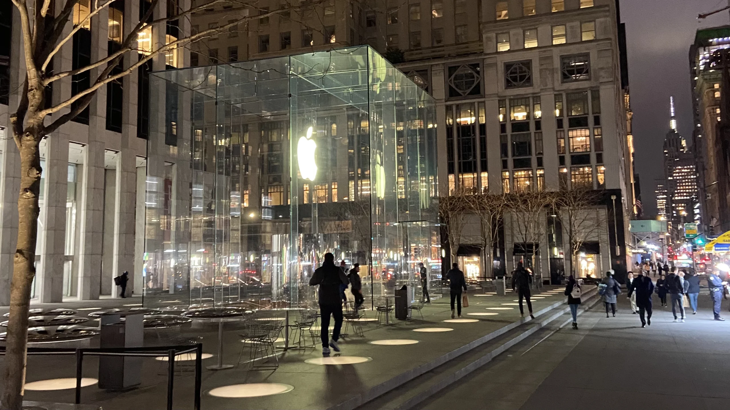 Pedestrians outside Apple’s illuminated glass cube entrance on Fifth Avenue in New York City at night.